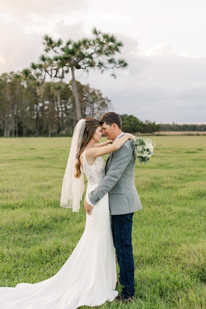 Newly wedded couple at the Heart D Farm after their outdoor ceremony. They are sharing an embrace in an open field with trees in the distance. Their foreheads are touching as they share an intimate moment.
