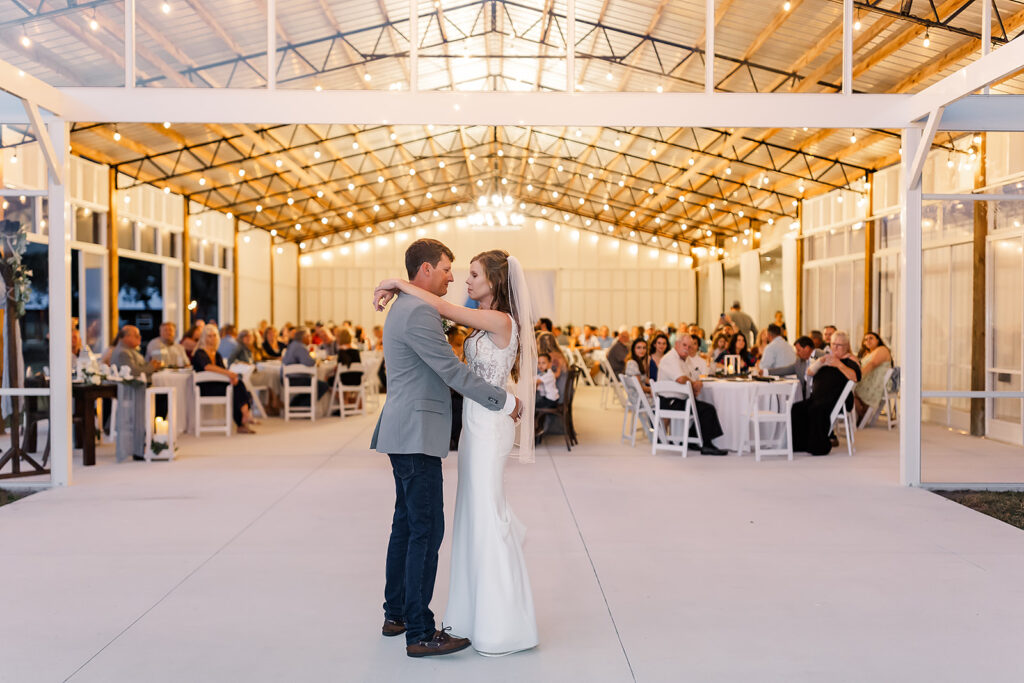 Newly wedded couple embrace during their first dance as husband and wife.  The are on the outdoor dance floor as evening is setting  in.  The wedding string lights glow and beging to transform the farm.  The clear glass wedding barn has the moving clear enclosure open and the gentle breezes are moving through to keep the guests comfortable.