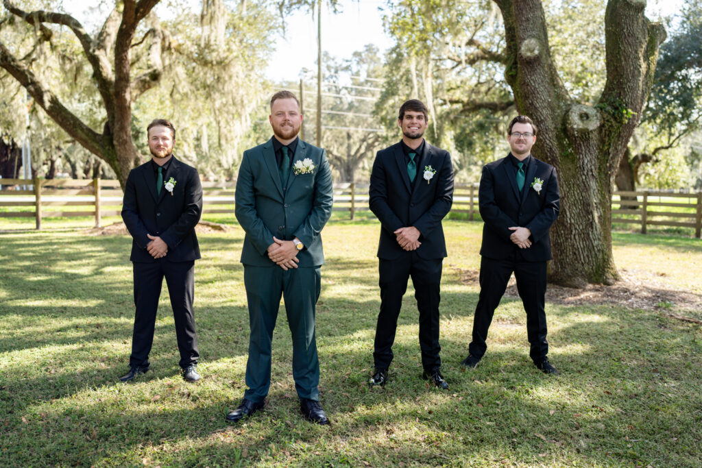 Groom and groomsmen stand at attention dressed in their finery. The groom is dressed in a dark green suit and all the groomsmen are looking sharp in black suits and dark green ties. Everyone is standing under the giant oaks.