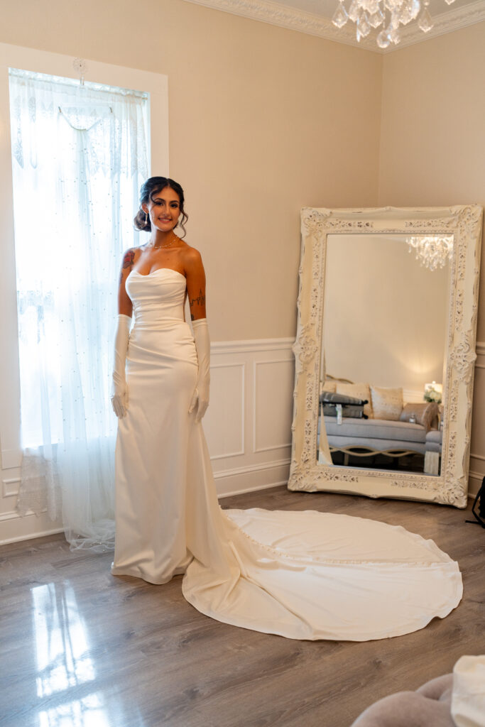 Gorgeous bride with dark coifed hair stands in front of the window with natural light and lace curtains in the bridal suite. Behind her is the full length white framed ornate leaning mirror. The walls are soft whites with a vintage vibe. The bride is wearing a fitted white shoulder-less wedding dress and long white gloves. Her train is encircling her feet. Part of the crystal chandelier can be seen in the high ceiling of the room.