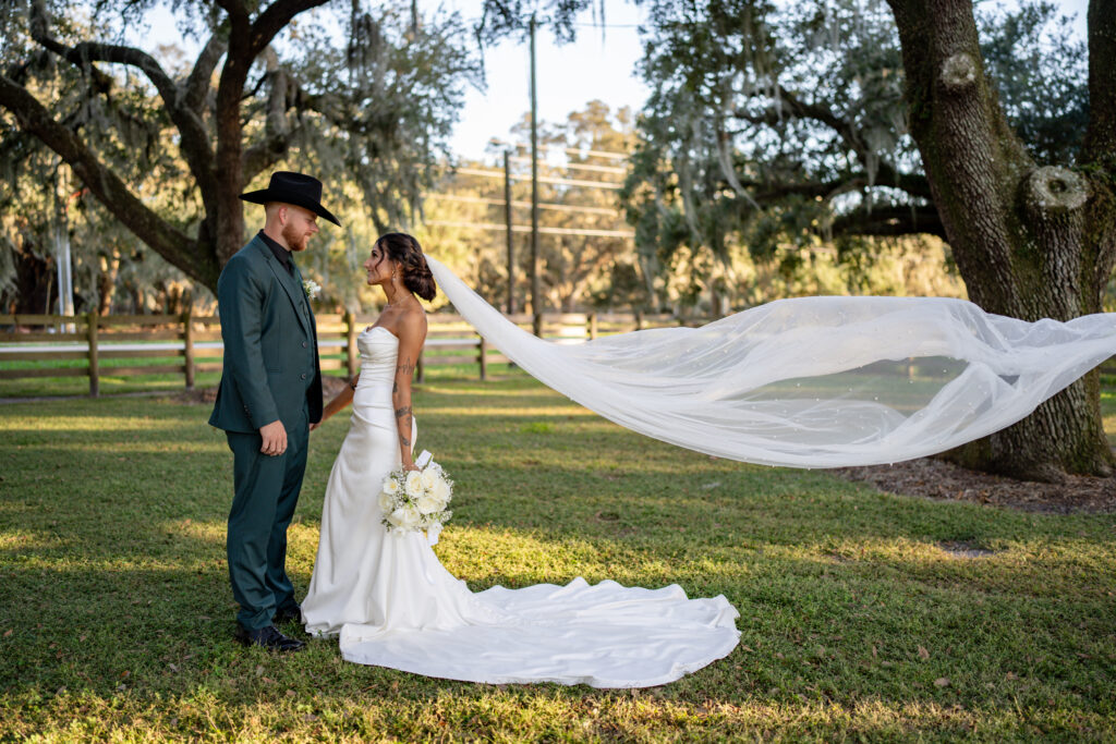 Stunning bride gazes into the eyes of her groom. She is stunning in her fitted white gown with long veil blowing horizontally in the breeze. A beautiful setting under the large oak trees.