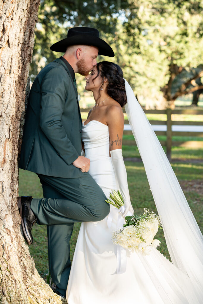 Bride and groom. Groom leans against a large oak tree and bride leans into him for a kiss on her forehead. They are under the huge oaks. He is wearing a dark green suit and a black cowboy hat. The bride is in a white fitted gown with a semi-opened back and open shoulders. She is wearing full length white gloves giving her an elegant look against the natural rustic background with a 4 board wood fence, pastures and towering Live Oak trees.