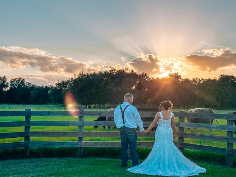 Recently wedded couple at the Heart D Farm in St. Cloud Florida after an outdoor ceremony stand holding hands facing a board fence and pasture while watching a gorgeous sunset.