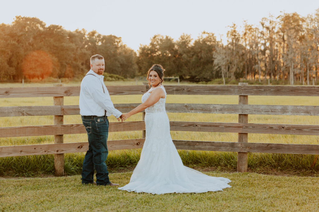 Beautiful couple on wedding day after the outdoor ceremony. Holding hands in front of a four board fence on a ranch in St. Cloud Florida.