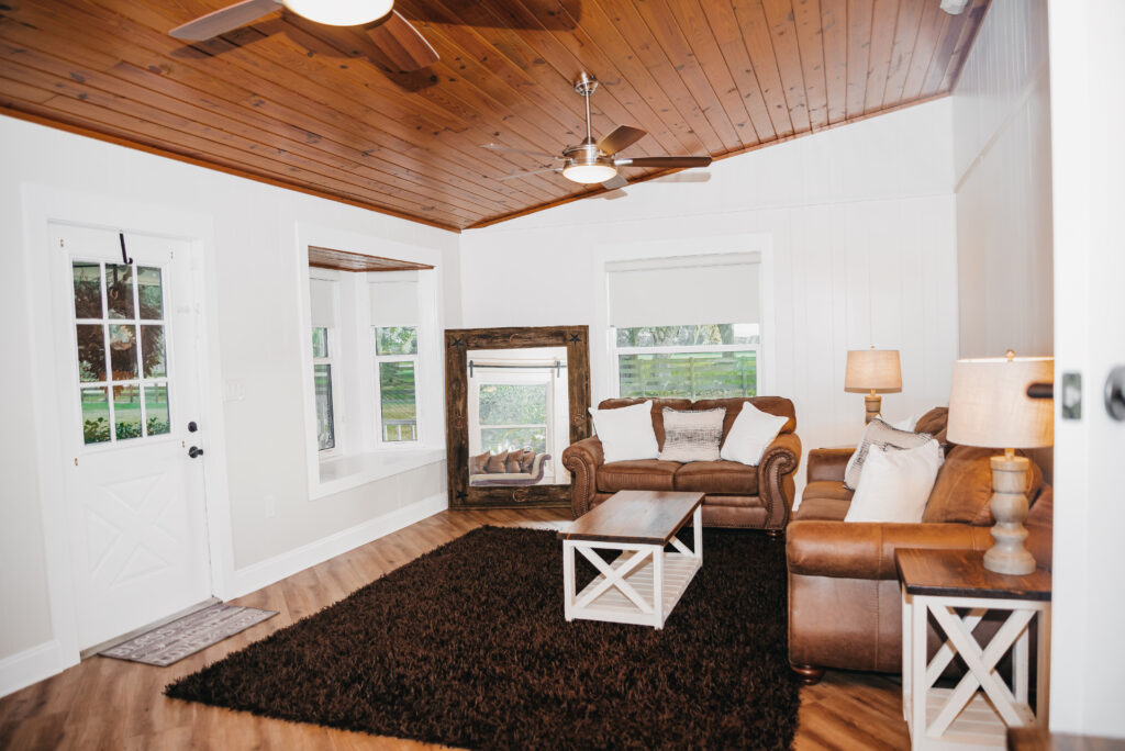 The Groom's Den is furnished plush brown sofas, a deep dark brown area rug and a rustic full length leaning mirror in the corner. The walls are done in soft whites and the vaulted ceiling is tongue groove with knotty pine stained wood. The sofa's have square white and gray pillows. The center table and end table are farm house white with stained wood tops. There are two lamps with gray wood bases and white shades. There is lots of natural light coming through the large windows and door with window.