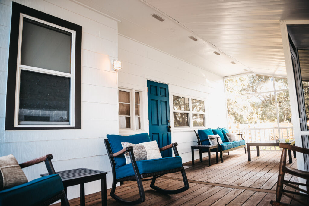 Warm and inviting screened back porch of the white farm house has wood floor and comfortable brown patio furniture and lovely blue cushions.  