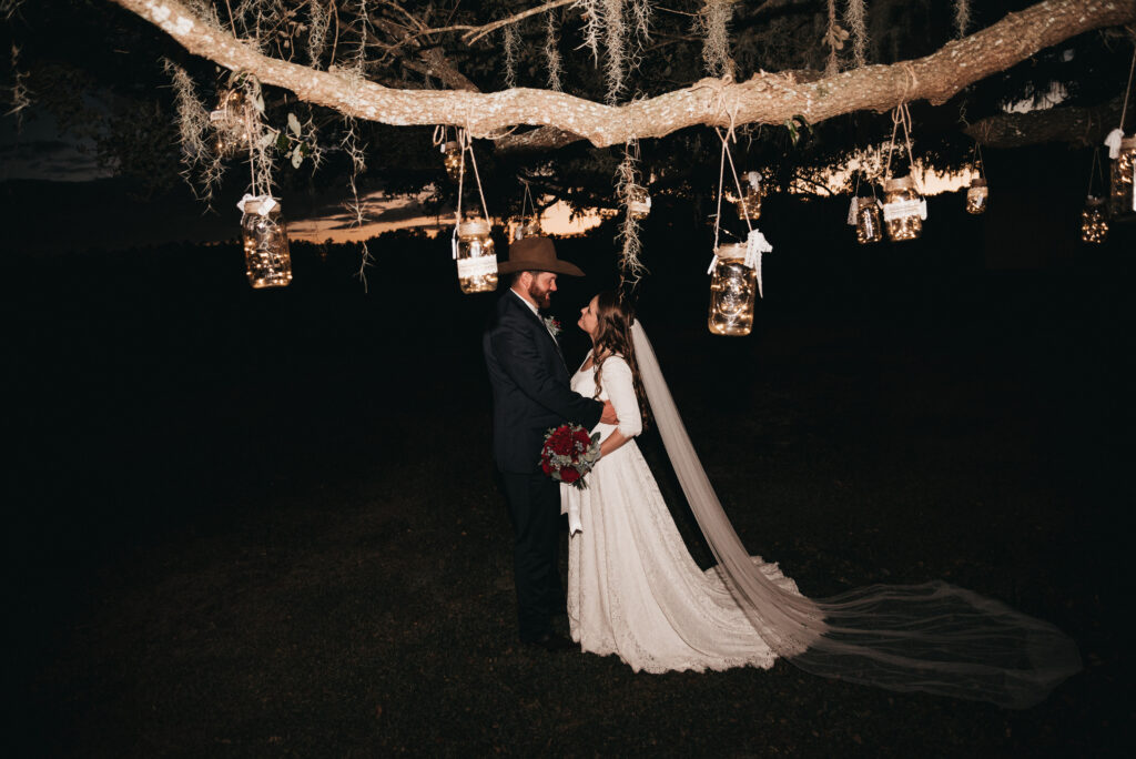 Recently wedded couple shares an embrace in the dark of night under the ancient oak trees with twinkling fairy lights in jars hanging from the limbs of the trees.