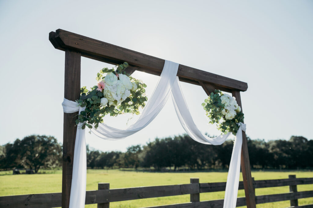 Photo is taken outside on the farm. It is the upper half of one of our traditional wood wedding arch which is decorated with white tulle, greenery and white flowers. The background is filled with large Live Oak trees in the distance, an open field and a four board fence. It is a bright sunny and cloud free day.