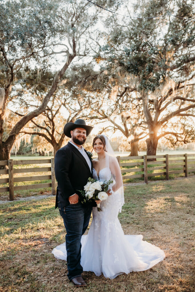 Lovely young couple are newly married and standing under the huge oak canopy with sunset filtering through the trees. The wood 4 board fence is in the background framing the open field behind them at the Heart D Farm Wedding Venue.