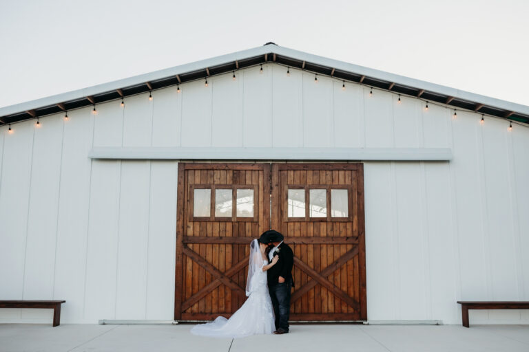 Newly wedded couple kiss in front of the large wood barn doors on the large white Big Barn pavilion. Fall wedding at Heart D Farm in Saint Cloud, FL