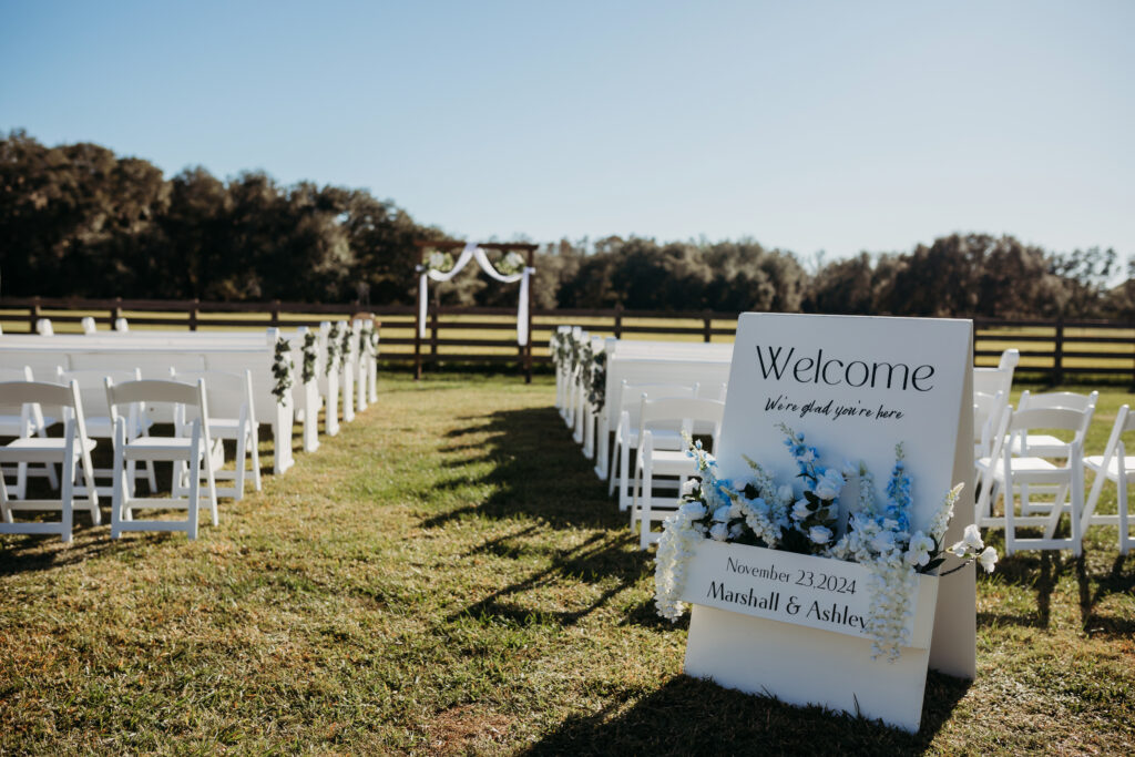 Outdoor ceremony site waiting for guests to arrive.  Overlooking an open pasture.  The traditional arch is decorated and the white church pews and white chairs are stunning against the lawn.  A welcome sign is at the head of the aisle and decorated with white and soft blue flowers. All at the Heart D Farm Wedding Venue in Kissimmee-St. Cloud Florida